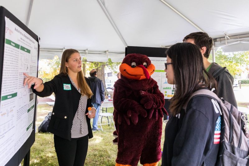 The HokieBird stands in the middle of a student presenter pointing towards their scientific research poster while two students pay close attention.