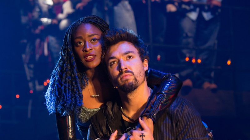  Two cast members of "Rent" face the camera. One is a Black woman with long braids parted in the middle with blue tips, wearing a grey tank top and black shrug. She stands behind a white man with brown hair and a brown beard and drapes one of her arms over his shoulder.