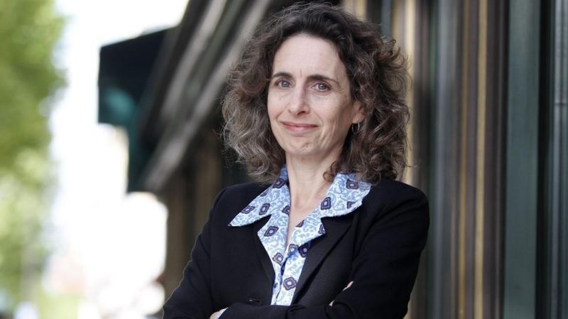  Author Elizabeth Kolbert, a middle aged white woman with curly, sandy brown hair and blue eyes, leans against the exterior wall of a building in this headshot. She wears a black blazer over a blue patterned button down shirt with a wide collar, and smiles towards the camera.