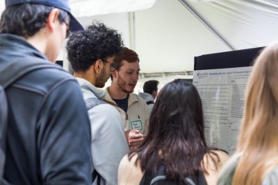 Through a crowd of four students, one student stands next to their scientific research poster outside under a tent and is talking to someone who asked them a question.