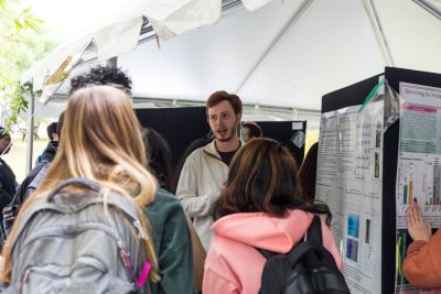 Through a crowd of students, one student stands next to their scientific research poster outside under a tent and is talking to someone who asked them a question.