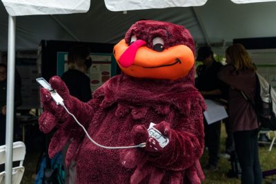 The HokieBird stands outside at an event holding a scientific instrument.