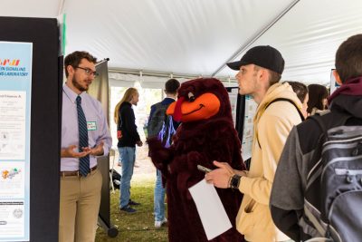 The HokieBird stands in the middle of a student presenter presenting their scientific research poster while another students pay close attention while taking notes on their phone.