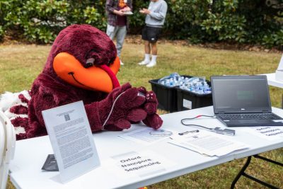 The HokieBird sits at a folding table holding a scientific instrument. The words "Oxford Nanopore Sequencing" are printed on an 11x14 white piece of paper and taped to the table. 