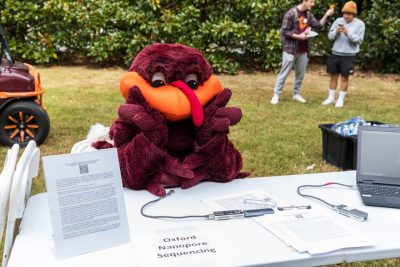 The HokieBird sits at a folding table and looks towards the camera. The words "Oxford Nanopore Sequencing" are printed on an 11x14 white piece of paper and taped to the table. 