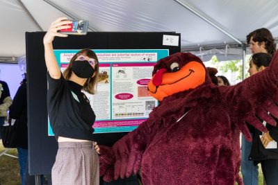 A student extends their arm to take a selfie with the HokieBird in front of their scientific research poster.
