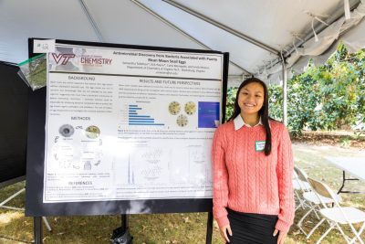 A student stands next to their scientific research poster outside under a tent in a symposium. 