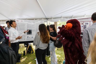 The HokieBird extends his arms in a V-shape towards a student presenter gesturing towards their scientific research poster. A crowd of students looking at the poster separate the HokieBird from the presenter.