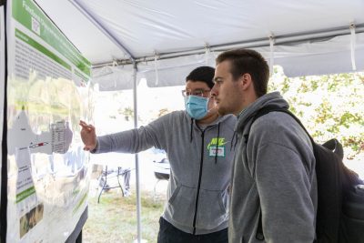 Two students look closely at a scientific research poster outside under a tent. One student is gesturing towards the poster.