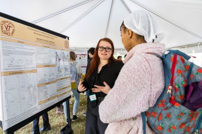 Two students stand in front of a scientific research poster. The student in the foreground is looking towards another student who is gesturing towards themselves as they talk.
