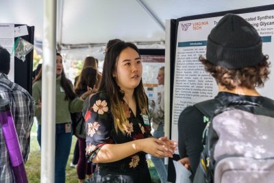 A student presenter talks with another student about their scientific research poster next to them outside under a tent.