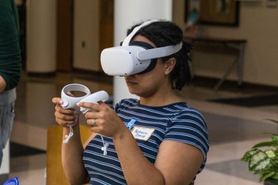 One student seated inside is wearing a virtual reality headset over their head and eyes and is holding onto hand controllers.