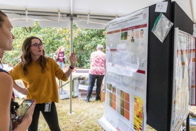Two students look towards a scientific research poster. One is gesturing towards their poster while the other is listening intently.