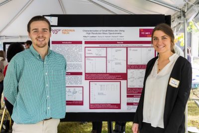 Two students stand next to their scientific research poster outside under a tent in a symposium. 