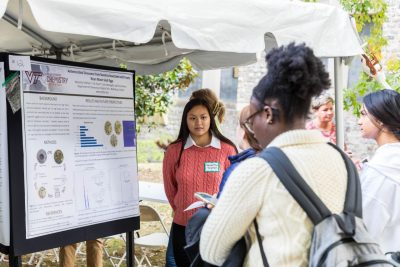 A student stands next to their scientific research poster outside under a tent in a symposium and listens intently to a student in front of a crowd.
