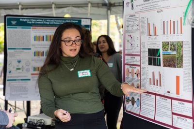 A student stands next to their scientific research poster outside under a tent in a symposium and points towards one of the boxes while in mid-dialogue.