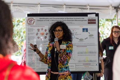 Biochemistry alumna holds a mic and PowerPoint clicker and gives a presentation to a crowd of students.