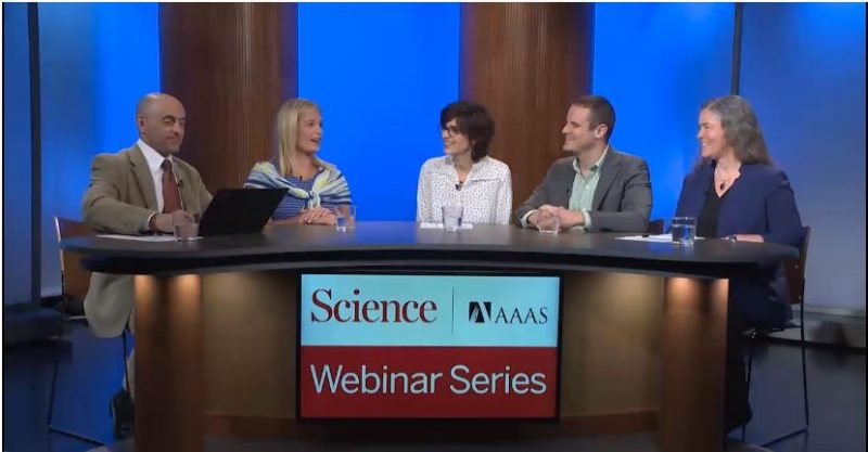 Five people sit at a news desk: a bald man in a brown suit and red tie, a blonde woman wearing a light blue shirt, a woman with short brown hair and a white shirt, a man in a grey suit and light blue shirt, and a woman with long blonde hair wearing dark blue. The sign on the desk reads “AAAS Science Webinar Series”