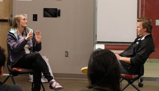 A blonde woman in a purple jacket and black pants sits across from a man with brown hair and a black jacket, explaining something.