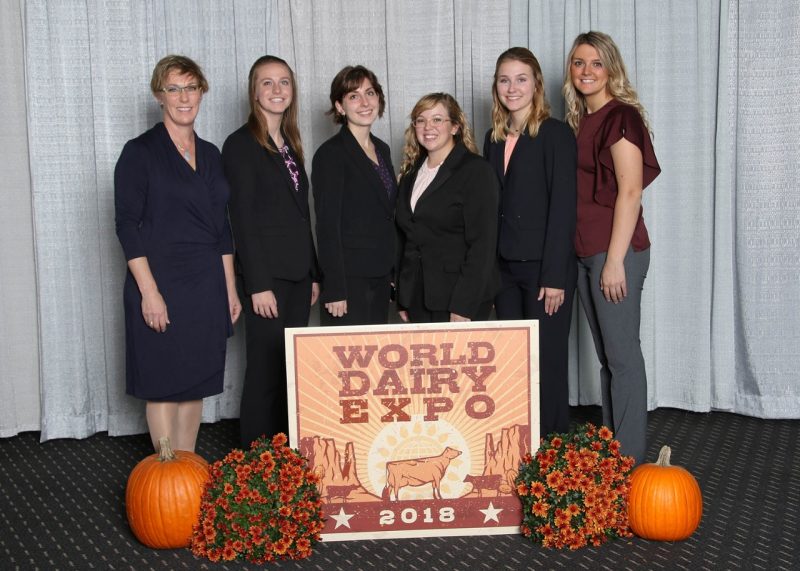 The 2018 VT Dairy Judging Team formal fall photo at the World Dairy Expo with autumn decorations of pumpkins and orange mums in the foreground.