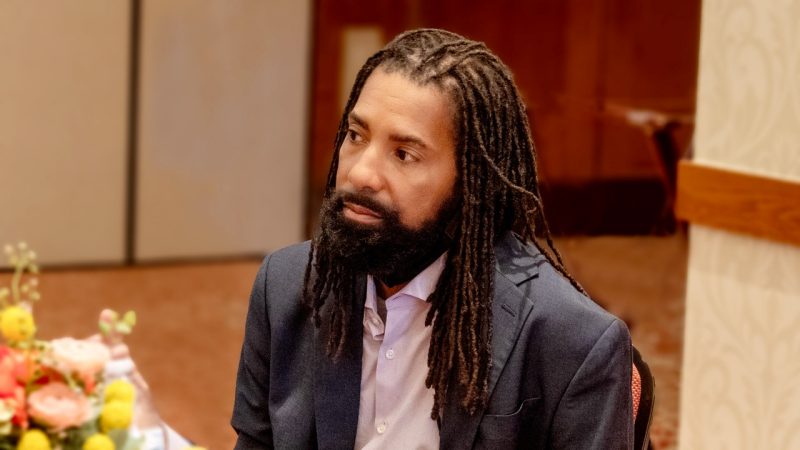 A Black male with long braids and a beard looks off to the left.