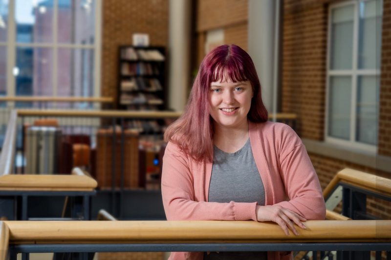 Millie wears a pink sweater, grey top, and has maroon-dyed hair. She smiles at the camera while leaning against a banister. A bookcase can be seen in the background.