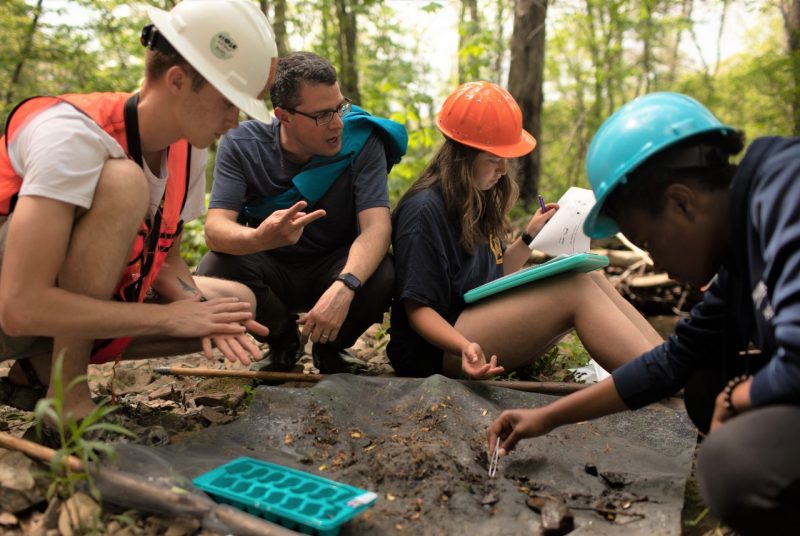 A group of people wearing hardhats collect samples from a net laid out on the ground. One person is kneeling and gesturing with his hands.