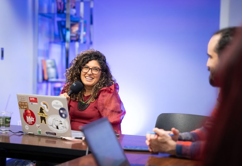 A smiling woman wearing a red shirt holds a microphone. She is sitting at a conference table in front of a laptop.