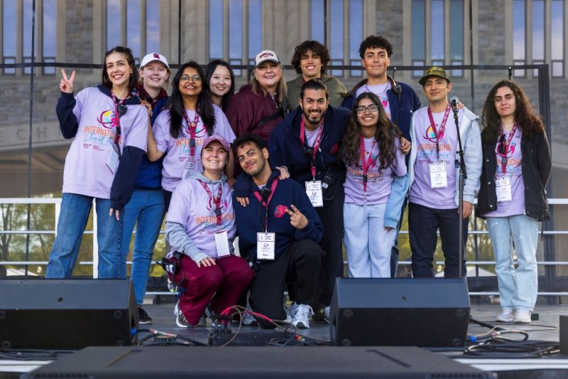 Several people, mostly wearing pink International Street Fair shirts, are standing together on stage posing for a group photo.