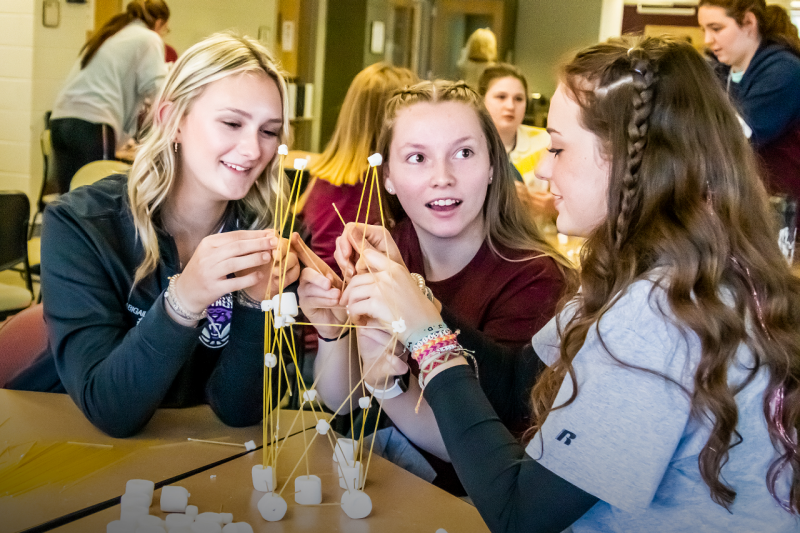 Three high school girls connect spaghetti noodles with marshmallows.