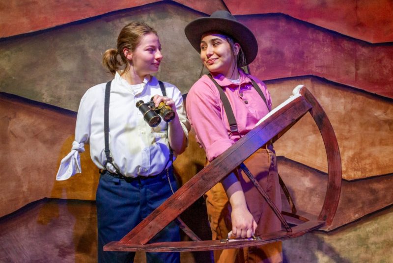 Two women, dressed as men, one with only one arm wearing a white shirt, blue jeans, suspenders and holding binoculars; the other wearing a hat, suspenders, red shirt, and holding a plane type of tool.