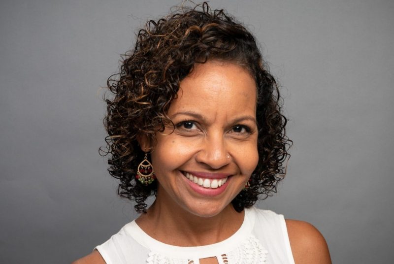 A woman with dark, short, curly hair, smiling, wearing a sleeveless white top